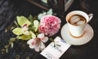a white cup of coffee sits on a table , accompanied by a bouquet of flowers and a welcome card at Mark Hotel Belgrade