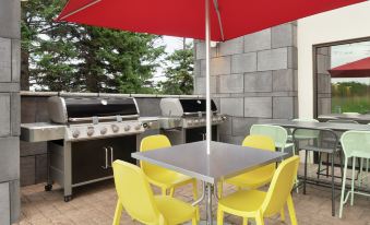 an outdoor dining area with a red umbrella , yellow chairs , and a grill in the background at Home2 Suites by Hilton Marysville