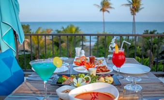 a table with various dishes and drinks , including a blue cocktail , is set up on a balcony overlooking the ocean at Seanery Beach Resort