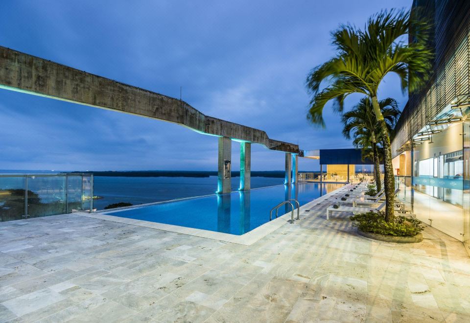 a large swimming pool is surrounded by palm trees and chairs , with a view of the ocean in the background at Cosmos Pacifico Hotel