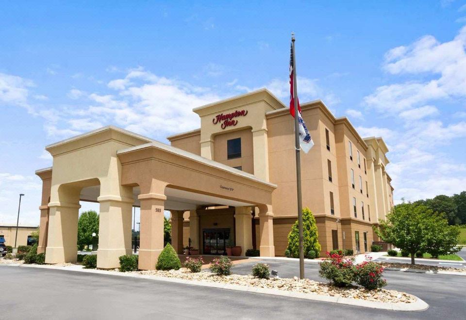 a large , beige - colored hampton inn hotel building with a flagpole in front , surrounded by trees and bushes at Hampton Inn Lenoir City