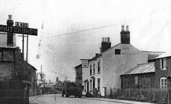 a black and white photo of a street with a building on the left side , a car driving down the road , and a bus parked in at The Sun Inn