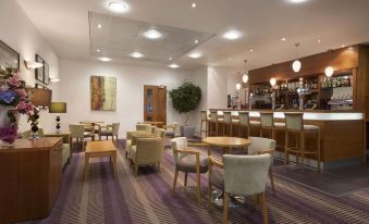 a modern lounge area with wooden tables , chairs , and a bar in the background , surrounded by white walls at Ramada Plaza by Wyndham Wrexham