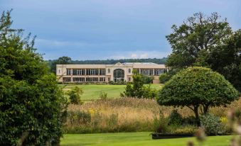 a large white building surrounded by green grass and trees , with a body of water in the background at Whittlebury Hall and Spa