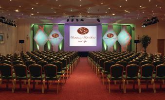 a large conference room with rows of chairs arranged in a semicircle , and a projector screen on the wall at Whittlebury Hall and Spa