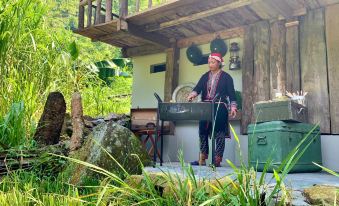 a man dressed in traditional chinese clothing is standing on a porch , holding a pan , surrounded by plants and a stone floor at Topas Riverside Lodge