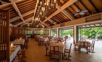 a large , open dining room with wooden tables and chairs , a chandelier hanging from the ceiling , and multiple pendant lights illuminating the space at The Residence Maldives at Dhigurah