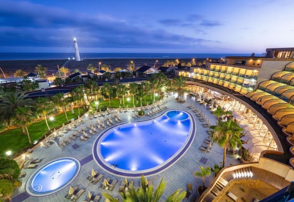 a large , blue swimming pool is surrounded by palm trees and lounge chairs in a resort with a city skyline in the background at Mur Faro Jandia Fuerteventura & Spa