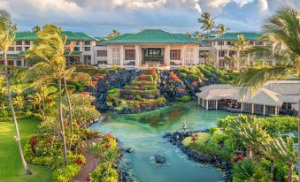 a large building with a green roof is surrounded by lush greenery and water , with people walking in the foreground at Grand Hyatt Kauai Resort and Spa