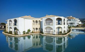 a large , white building with multiple balconies and arches is reflected in the water of a swimming pool at Mitsis Selection Laguna