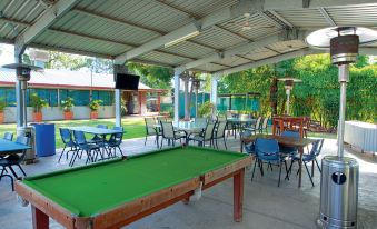a green pool table in a covered outdoor area , surrounded by chairs and tables , with a swimming pool visible in the background at Discovery Parks – Biloela
