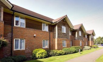 a brick building with multiple windows and a red roof is surrounded by greenery , bushes , and trees at Premier Inn Tewkesbury