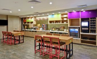 a large , modern cafeteria with wooden tables and chairs , a counter , and a variety of food items on display at Home2 Suites by Hilton Mechanicsburg