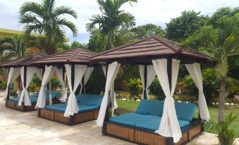 a row of blue lounge chairs under a wooden canopy , surrounded by palm trees and greenery at Oasis Resort