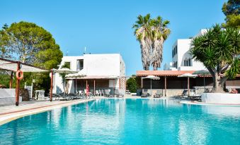 a large outdoor swimming pool surrounded by a hotel , with several lounge chairs and umbrellas placed around the pool area at Anamar Patmos