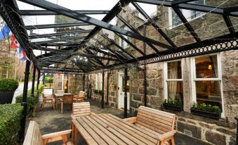 a wooden table and benches under a black - metal structure , with stone buildings in the background at Loch Kinord Hotel