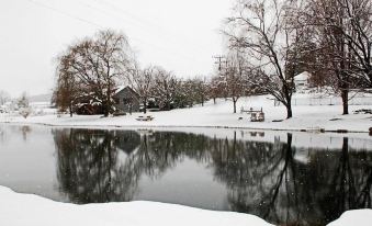 a serene winter scene of a pond surrounded by snow - covered trees , with a small house in the background at Steeles Tavern Manor