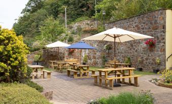 a garden with wooden benches and umbrellas , set against a stone wall with trees and greenery at The Celtic Royal Hotel
