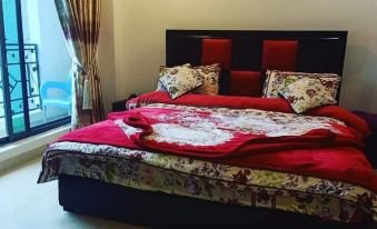 The bedroom is furnished with two beds adorned with red pillows, a side table featuring a lamp and a clock, and a window dressed with curtains at Taj Hotel and Restaurant