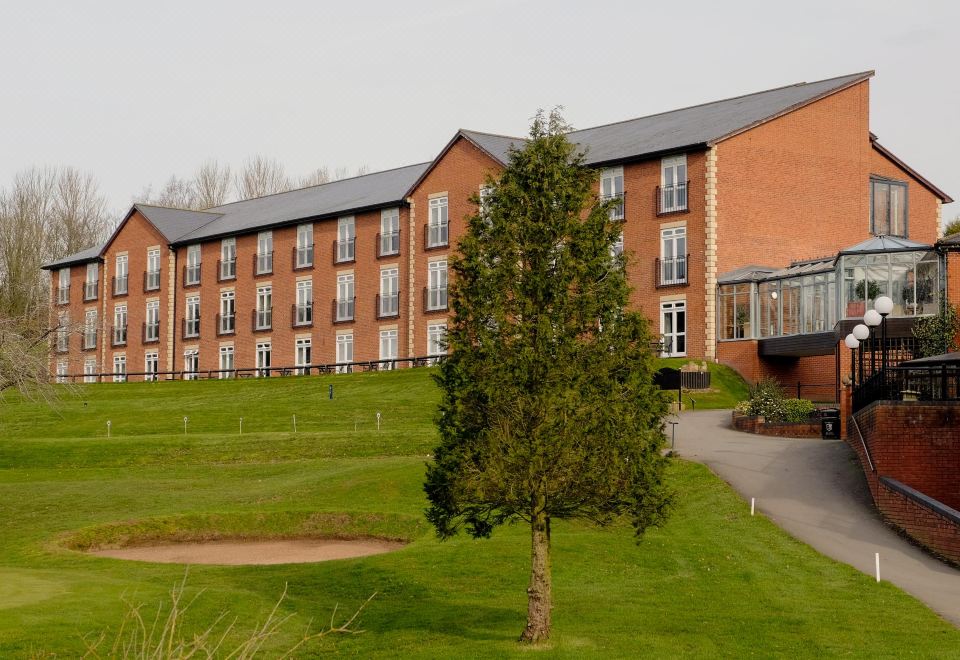 a large brick building surrounded by a golf course , with a tree in the foreground at Macdonald Hill Valley Hotel, Golf and Spa