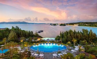 a large outdoor pool with a water slide is surrounded by lush greenery and overlooks a body of water at Halekulani Okinawa