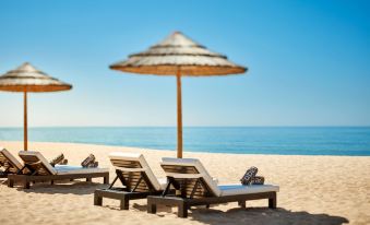 a row of lounge chairs under an umbrella on a sandy beach , with the ocean in the background at Tivoli Marina Vilamoura