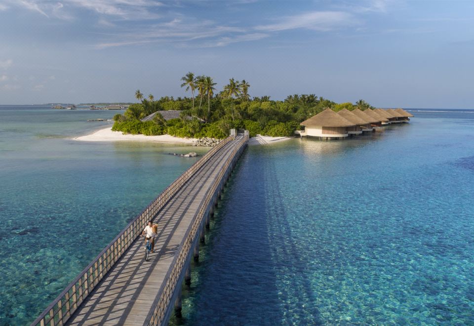 a man riding a bicycle across a wooden bridge over the ocean , with palm trees in the background at The Residence Maldives at Dhigurah