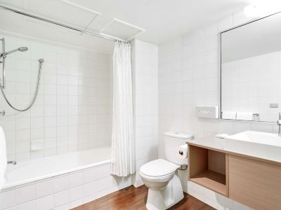 a modern bathroom with white tiles , wooden flooring , and a toilet , as well as a sink and bathtub at Ibis Melbourne Hotel and Apartments