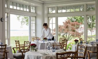 a woman in a white robe standing near a dining table with chairs and a large window at Inn at Perry Cabin