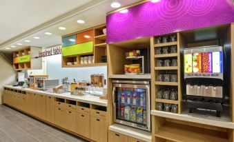 a modern cafeteria with wooden cabinets , a purple wall , and a counter equipped with various beverages and snacks at Home2 Suites by Hilton Helena