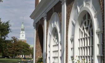 a white brick building with arched windows , surrounded by greenery and a tall steeple in the background at Hanover Inn Dartmouth