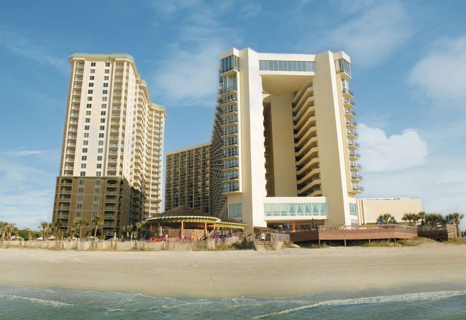 a large building is situated on the beach , surrounded by tall buildings and a sandy shoreline at Hilton Myrtle Beach Resort