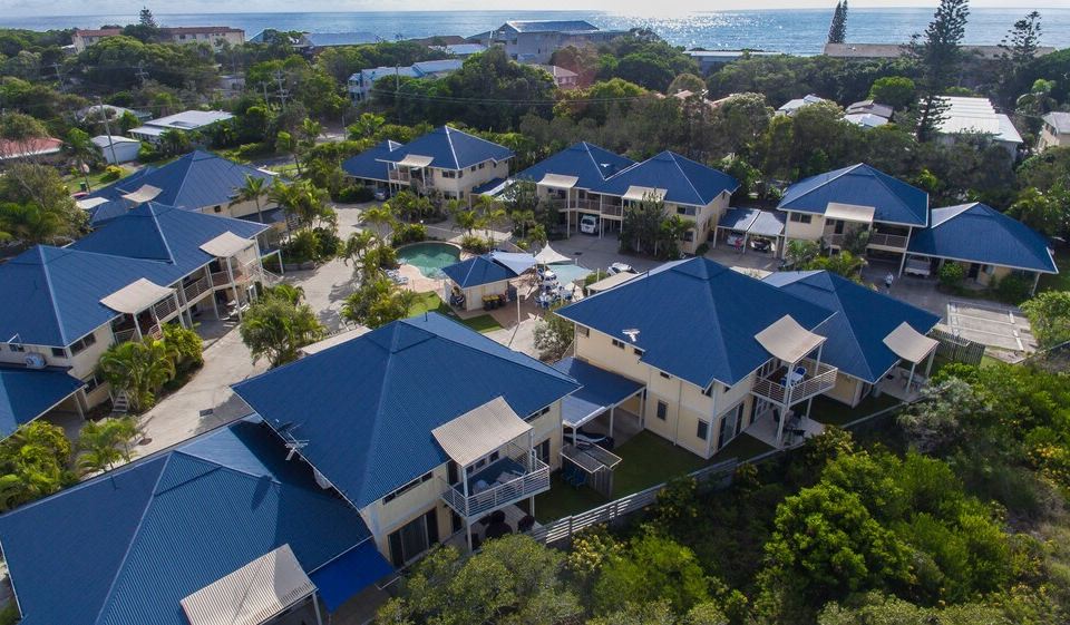aerial view of a row of blue - roofed houses surrounded by trees , with the ocean visible in the background at Hastings Cove Holiday Apartments