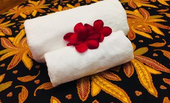 two white towels are neatly folded on a bed with red flowers in between , set against a decorative floral patterned tablecloth at Kampong Pinang Sebatang