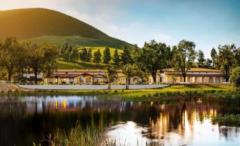 a serene landscape with a lake , trees , and buildings , reflecting the sun 's rays on the water 's surface at La Lomita Ranch