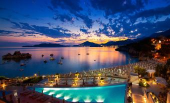 a large outdoor pool is surrounded by lounge chairs and lit up at night , with boats in the water and mountains in the background at Maestral Resort & Casino