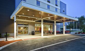 a modern , white - washed building with a covered entrance and seating areas , lit up at night , under a blue sky at Home2 Suites by Hilton Walpole Foxboro