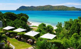 a beautiful tropical beach with clear blue water , white umbrellas , and green trees , set against the backdrop of mountains and clear skies at Raffles Seychelles