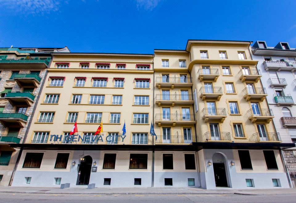 a large yellow building with multiple flags flying in front of it , surrounded by trees and other buildings at Hotel Geneva by Fassbind