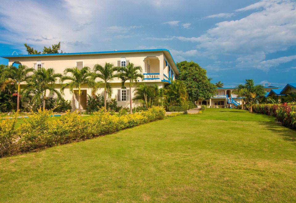 a large white building with a blue roof , surrounded by green grass and palm trees , under a cloudy sky at Travellers Beach Resort