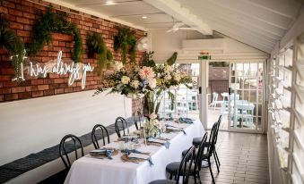 a long dining table with white tablecloths and chairs , surrounded by a brick wall and greenery at The Clan Terrigal
