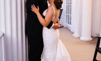 a newlywed couple in wedding attire embracing each other on a porch with columns and a white building in the background at Avon Inn