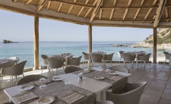 a restaurant with a thatched roof and large windows overlooking the ocean , featuring multiple dining tables and chairs at Falkensteiner Resort Capo Boi