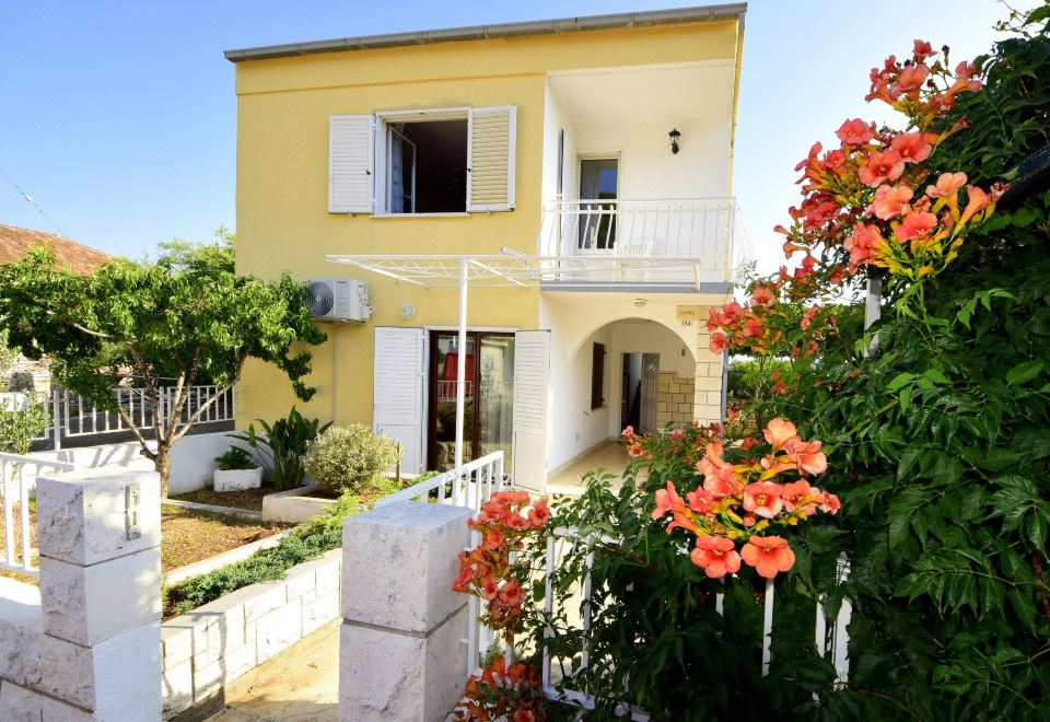 a yellow house with a white balcony and red flowers in front of it , surrounded by trees and plants at Lolita