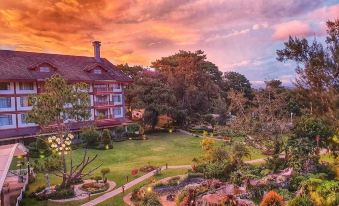 a large house is surrounded by a well - maintained garden with trees and bushes , as the sun sets in the background at The Manor at Camp John Hay