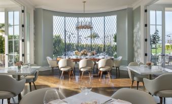 a modern dining room with a large window , a chandelier , and multiple tables set for dinner at Rosewood Miramar Beach