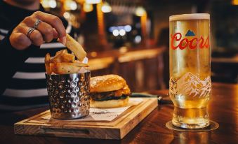 a person is holding a glass of beer and sitting at a table with a burger and fries at The Knaresborough Inn - the Inn Collection Group