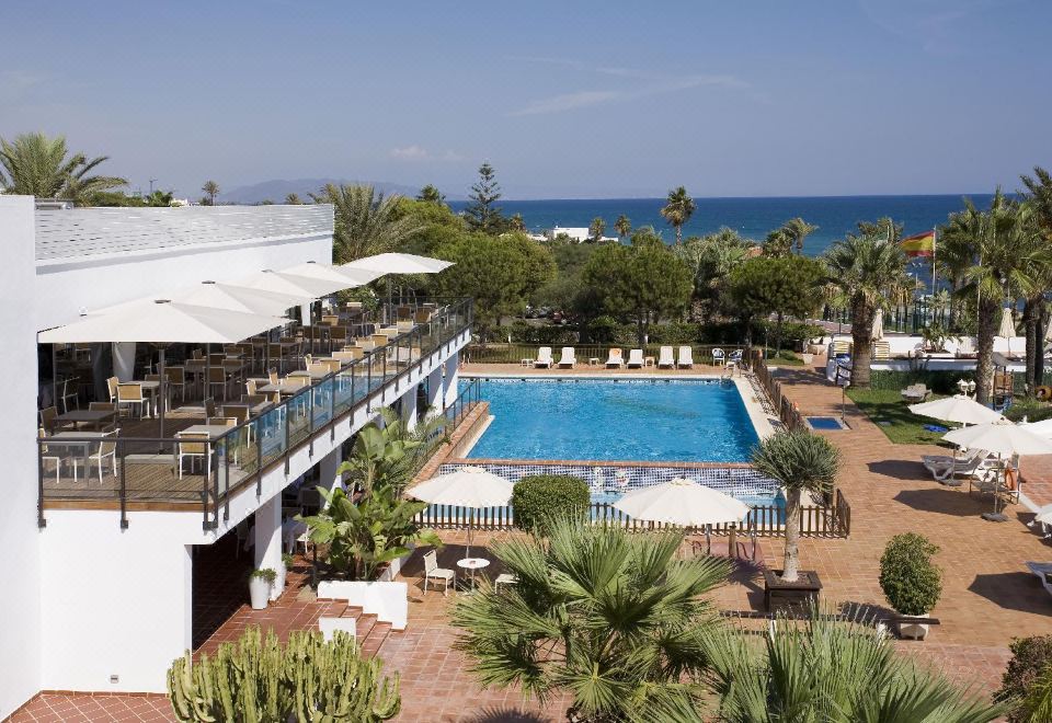 a beautiful resort with a large swimming pool , umbrellas , and palm trees , situated near the ocean at Parador de Mojacar