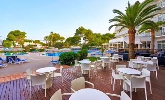 a poolside area with white tables and chairs , surrounded by palm trees and a swimming pool at Globales Cala Blanca