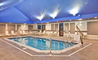 an indoor swimming pool with a blue ceiling , surrounded by white lounge chairs and umbrellas , and a row of chairs on the edge of the pool at Homewood Suites by Hilton Dover - Rockaway